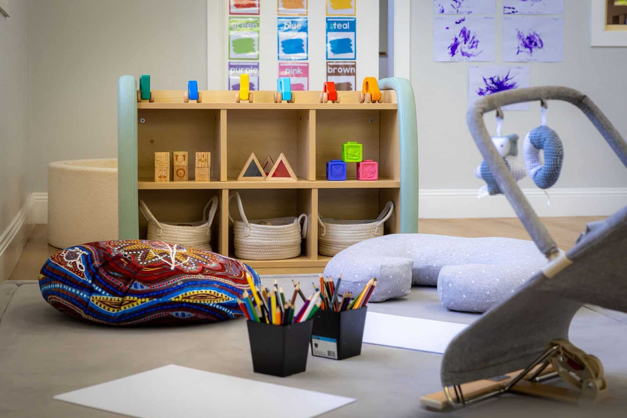 Nursery play corner with low timber shelves, soft cushions and a baby bouncer